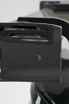 Front close-up of a black BMW G87 M2 featuring an aggressive carbon fiber front bumper, deep front splitter, large air intakes, and gloss carbon fiber aero details in a studio setting.