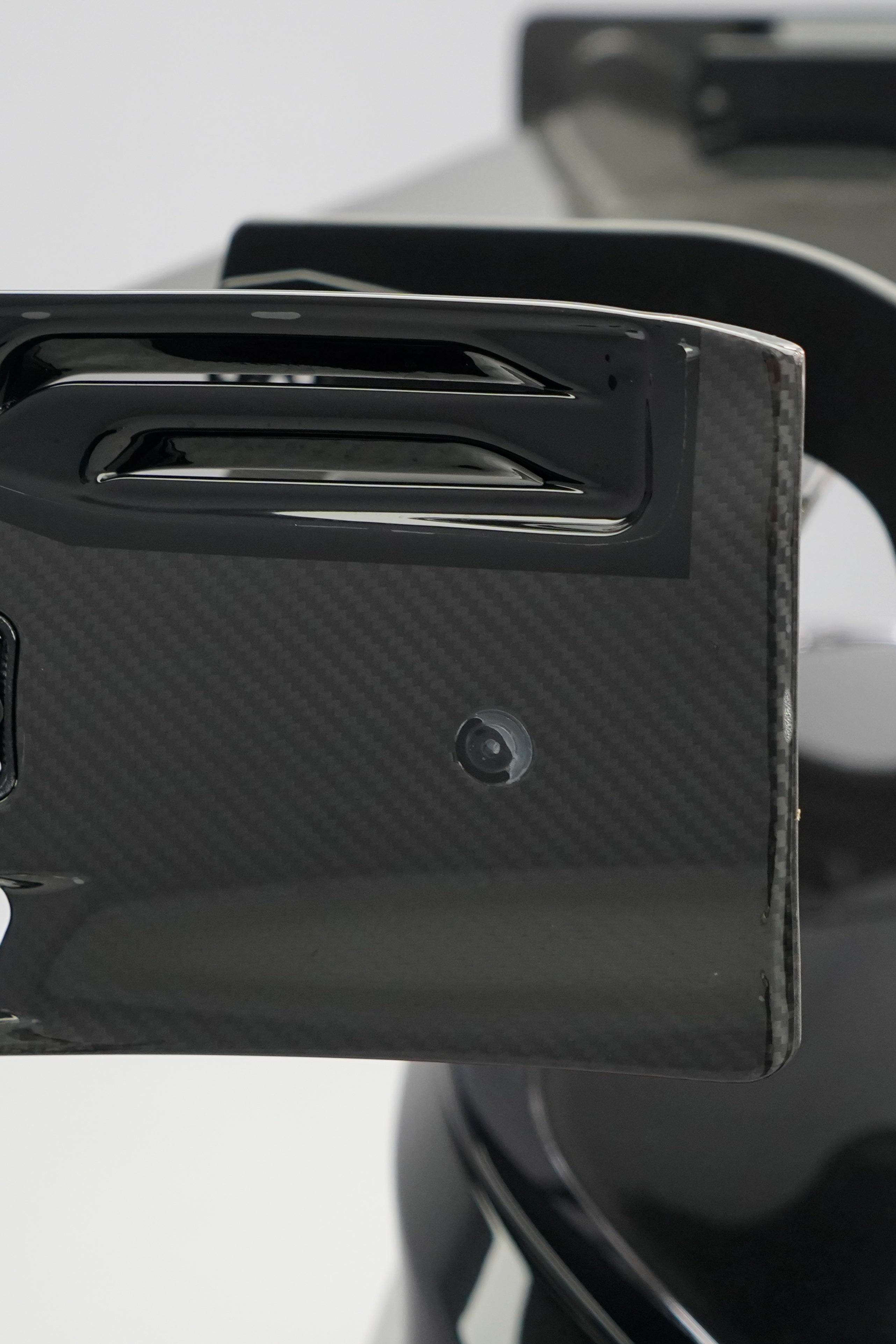 Front close-up of a black BMW G87 M2 featuring an aggressive carbon fiber front bumper, deep front splitter, large air intakes, and gloss carbon fiber aero details in a studio setting.