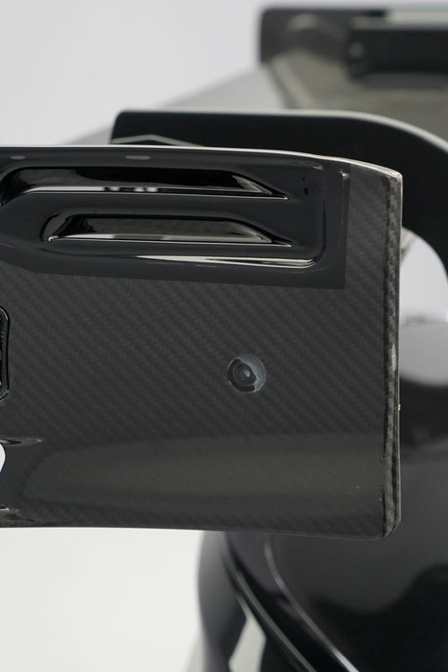 Front close-up of a black BMW G87 M2 featuring an aggressive carbon fiber front bumper, deep front splitter, large air intakes, and gloss carbon fiber aero details in a studio setting.