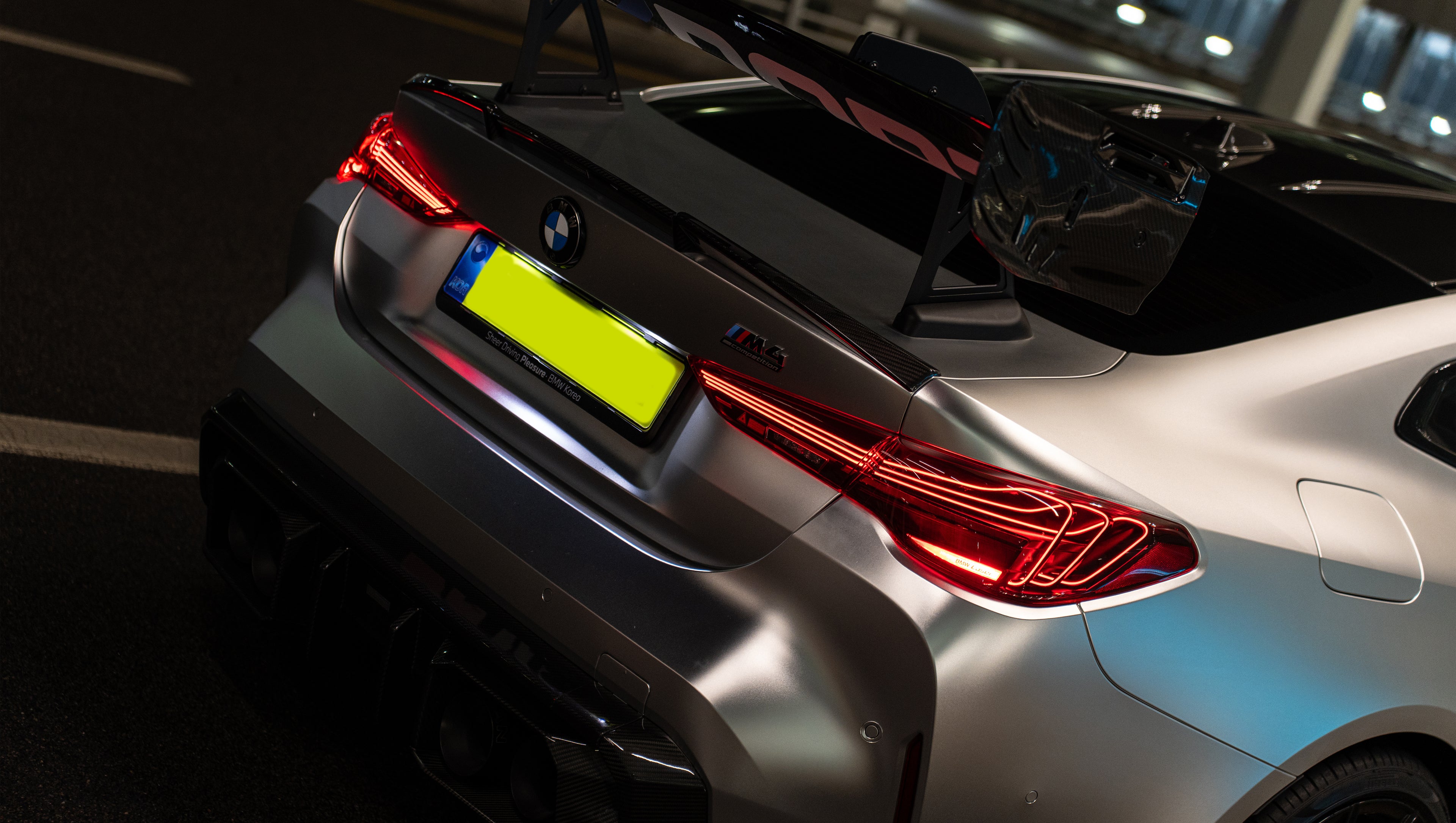 Matte gray BMW G82 M4 featuring an aggressive REVOZPORT carbon fiber aero kit with vented hood, front canards, wide fender vents, and illuminated LED headlights in a nighttime parking garage.