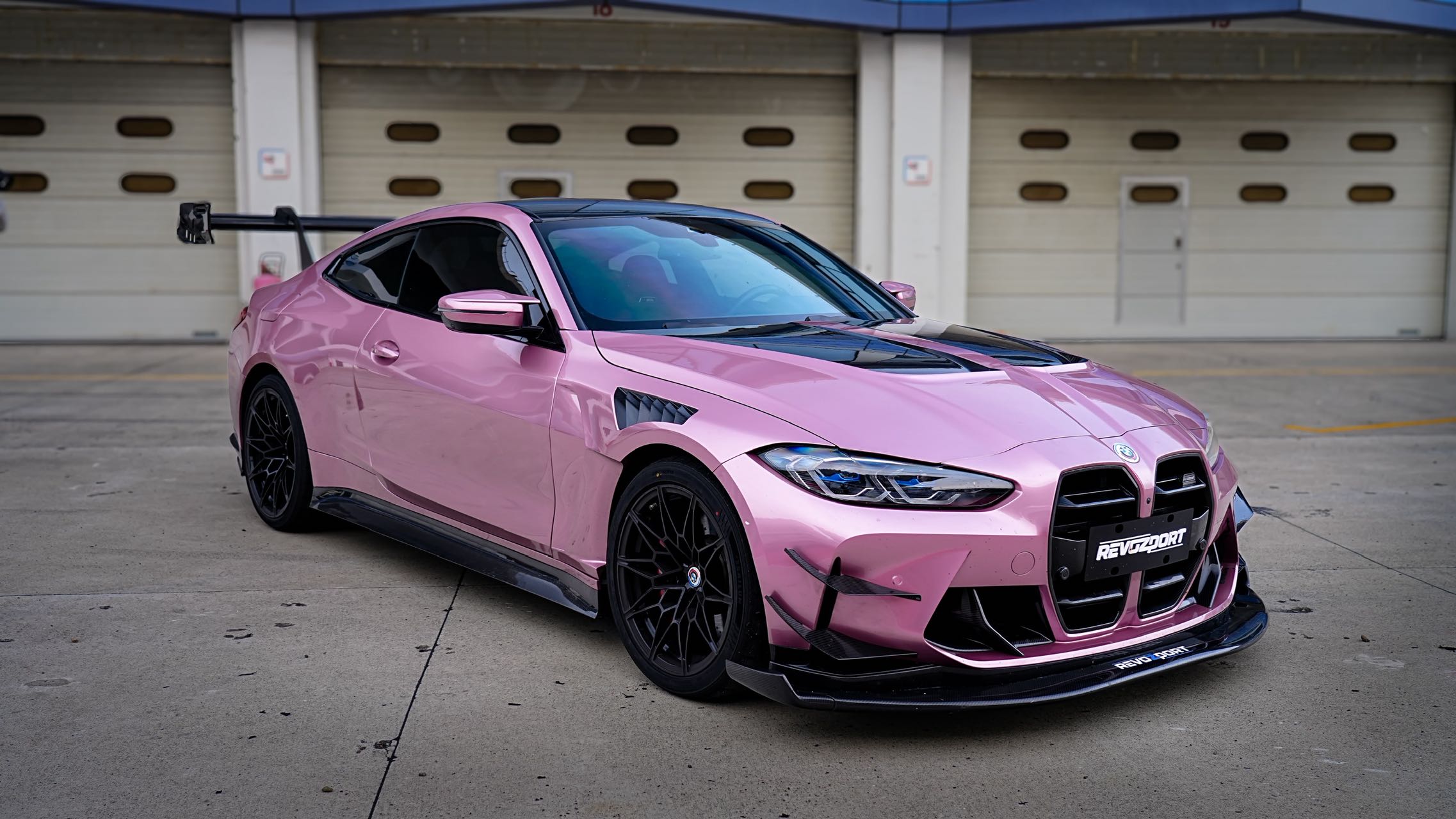 Pink BMW G82 M4 fitted with a REVOZPORT carbon fiber widebody aero kit, front splitter, hood vents, and side skirts on a race track pit lane.