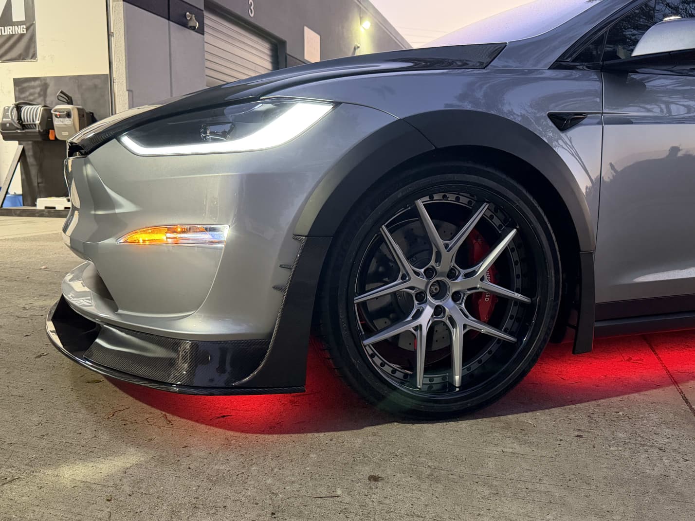 Tesla Model X featuring an aggressive REVOZPORT carbon fiber body kit with front lip, vented hood, wide fender extensions, side skirts, and illuminated red underglow lighting at dusk.