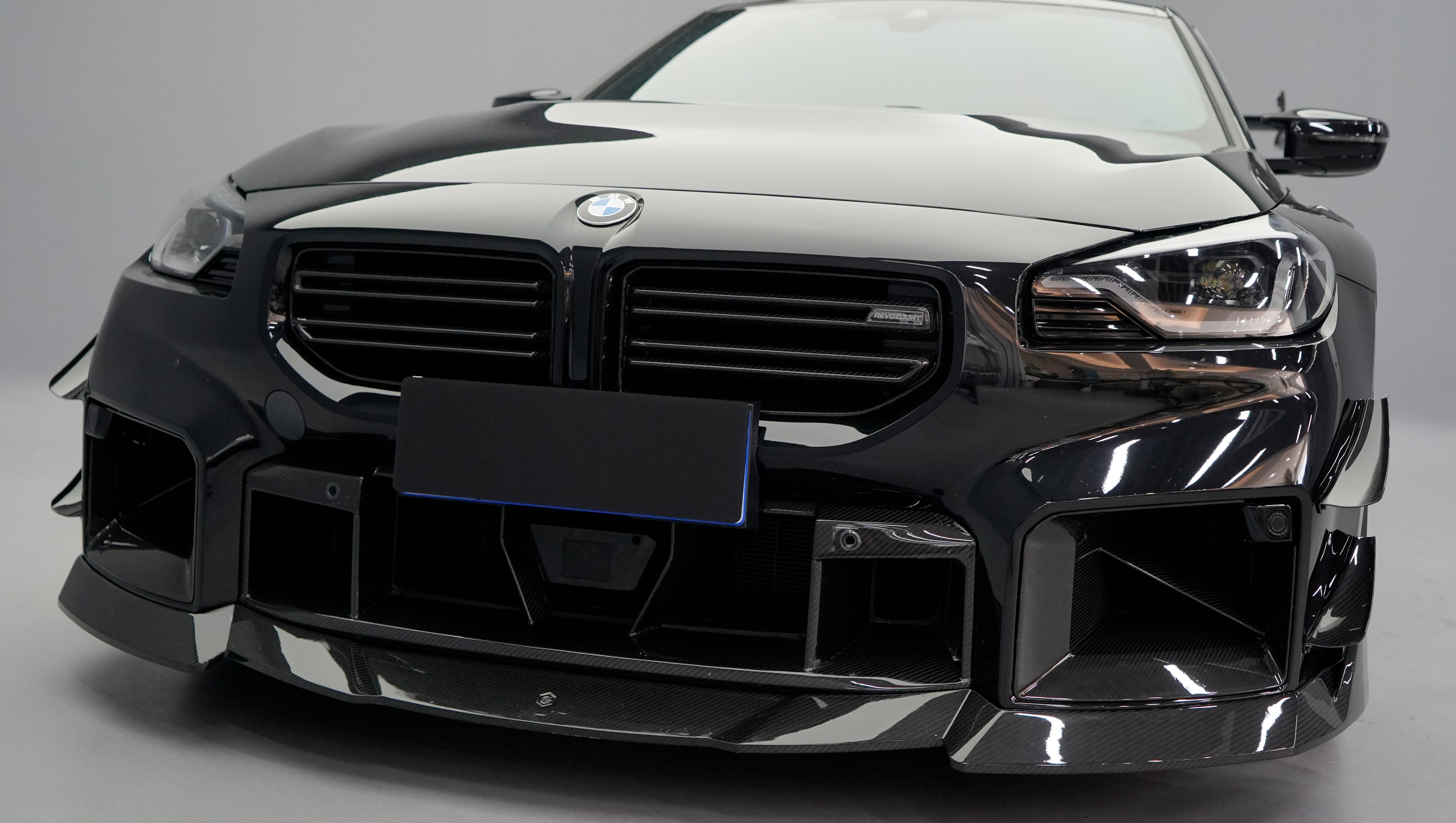 Front close-up of a black BMW G87 M2 featuring an aggressive carbon fiber front bumper, deep front splitter, large air intakes, and gloss carbon fiber aero details in a studio setting.