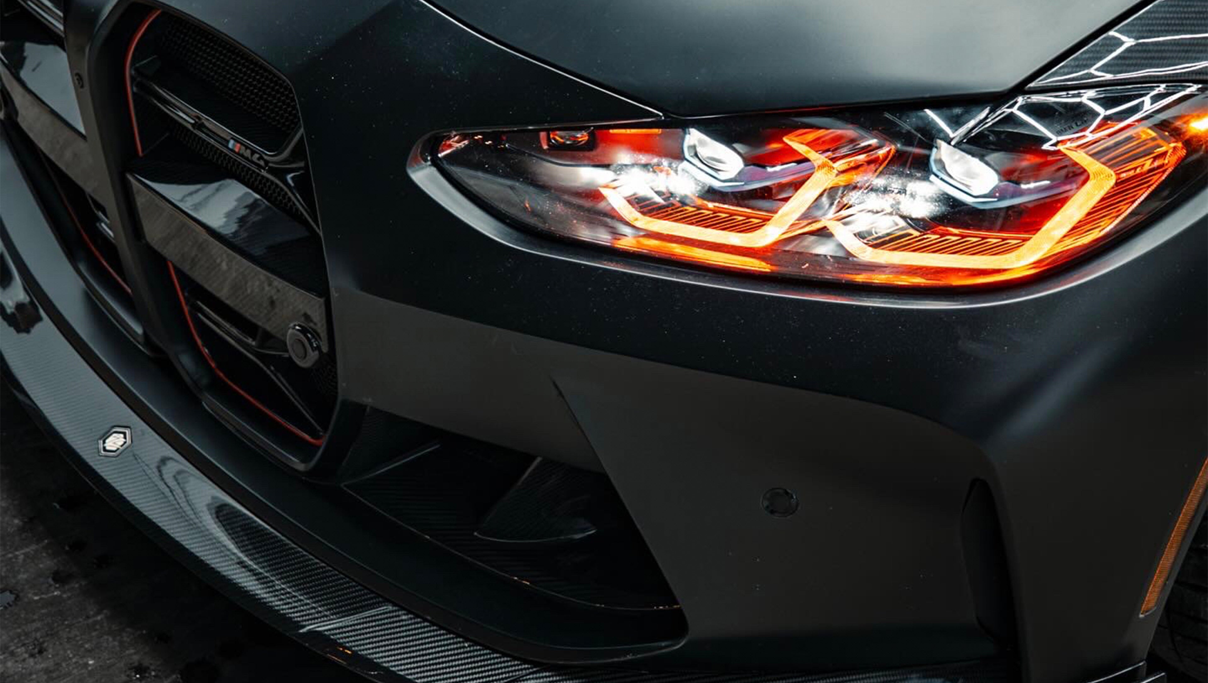 Matte black BMW G82 M4 with carbon fiber front splitter, side skirts, vented hood, bronze wheels, and oversized rear wing parked on a city street at night with headlights on.