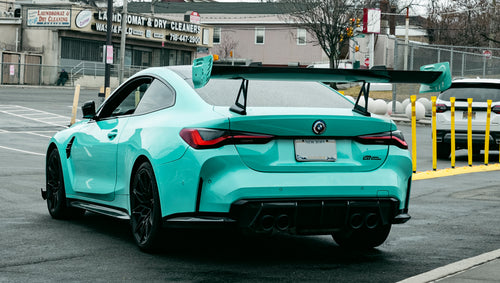 Mint green BMW G82 M4 with carbon fiber front splitter, side skirts, and large rear wing parked on a wet urban street outside a modern building.