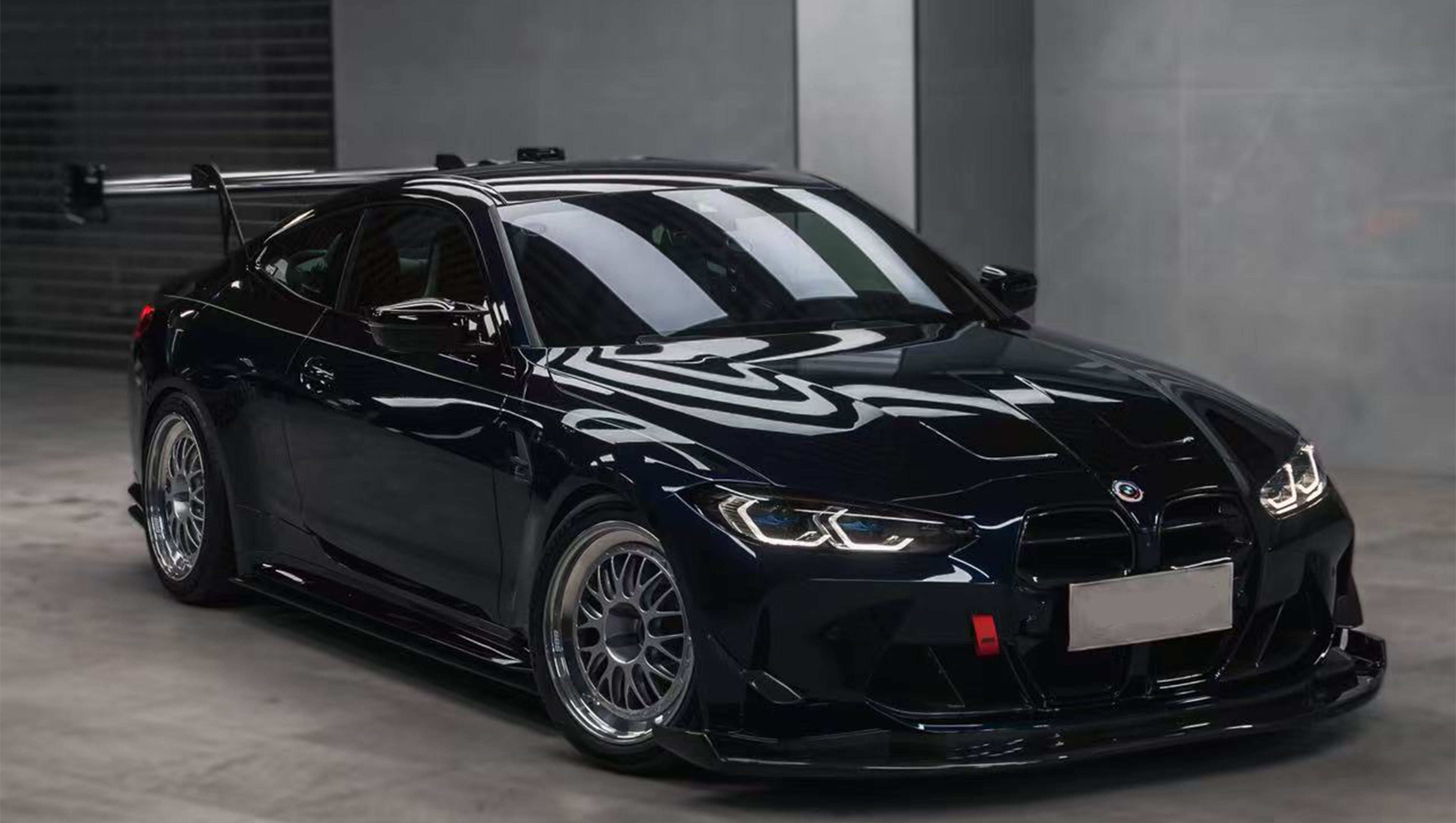 Dark blue BMW G82 M4 equipped with carbon fiber front splitter, side skirts, vented fenders, and a large rear wing, parked inside a modern luxury garage.