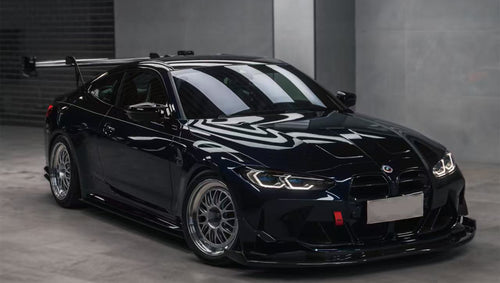 Dark blue BMW G82 M4 equipped with carbon fiber front splitter, side skirts, vented fenders, and a large rear wing, parked inside a modern luxury garage.