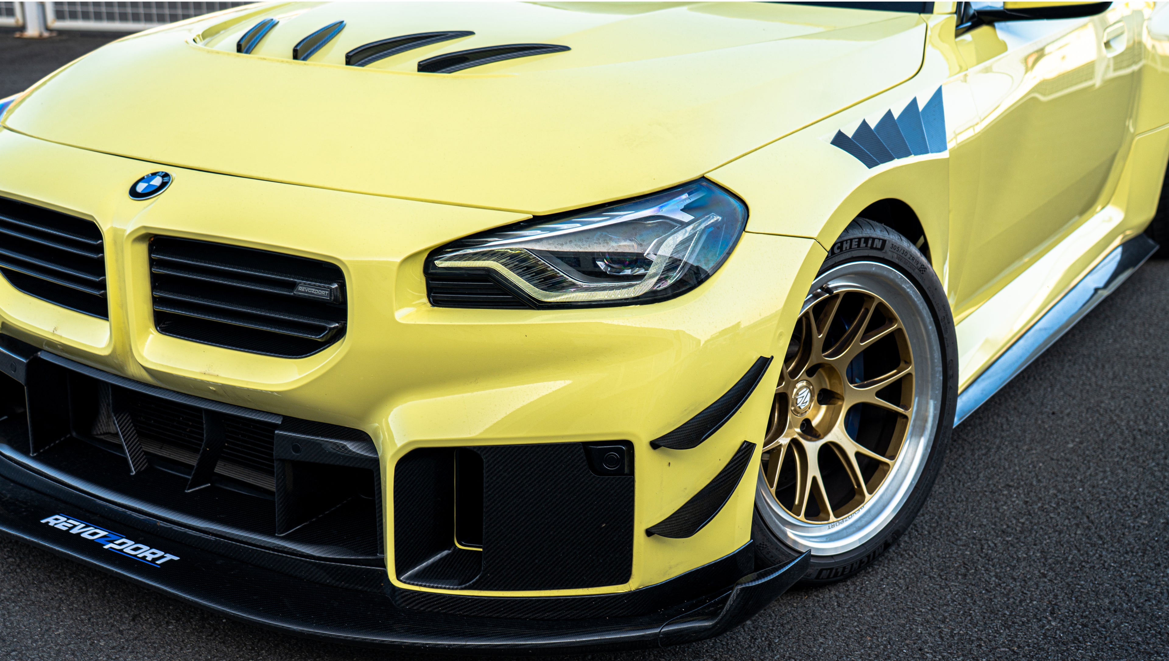 Pastel yellow BMW G82 M4 with carbon fiber front splitter, hood vents, side canards, bronze wheels, and large rear wing parked at a race track facility during golden hour.