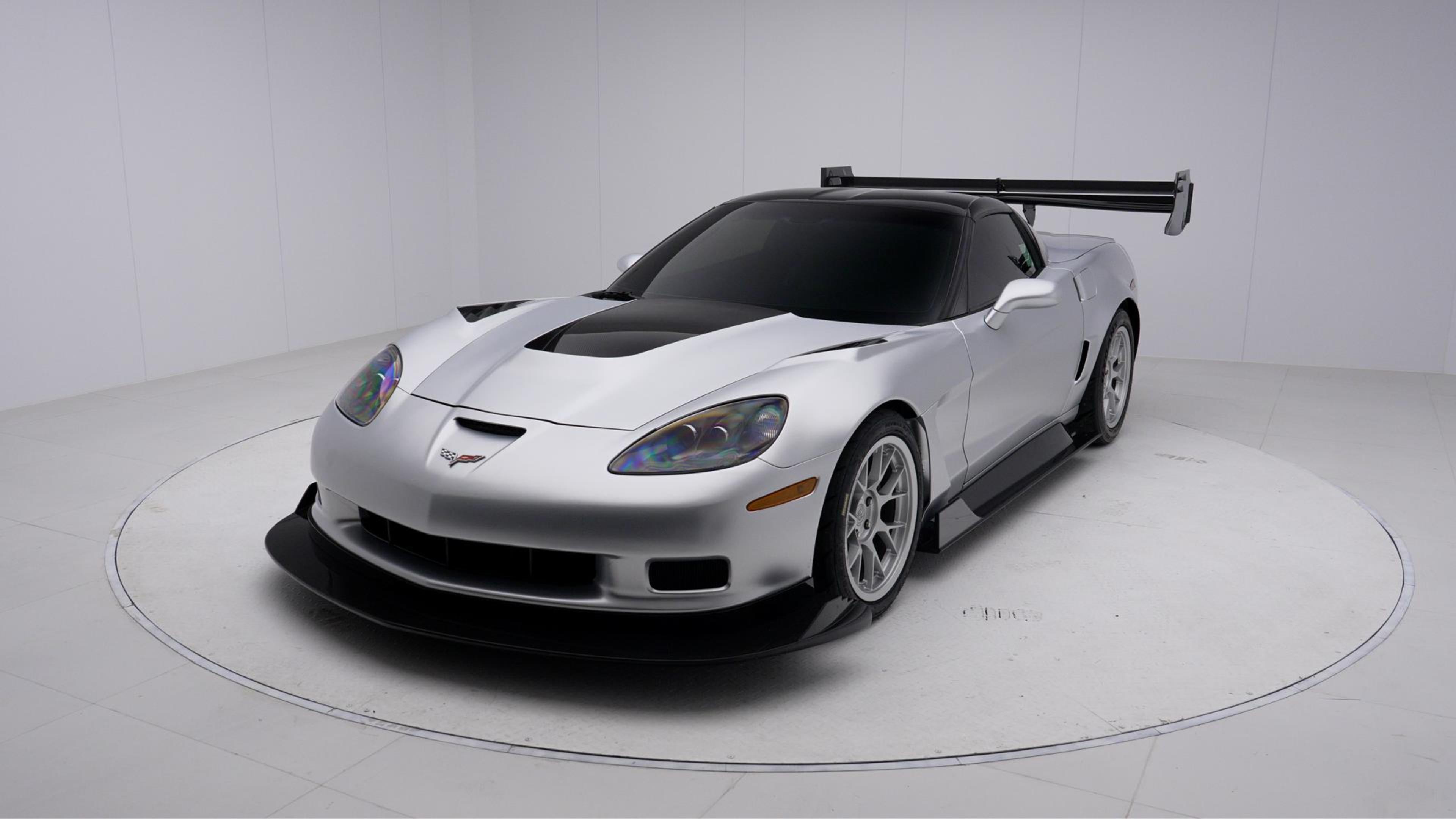 Close-up of a silver wide-body Chevrolet Corvette performance car displayed indoors, featuring aggressive aerodynamic bodywork, carbon fiber front splitter, vented hood, and lightweight multi-spoke wheels. The image highlights the custom motorsport styling and premium carbon fiber aero components under exhibition lighting.