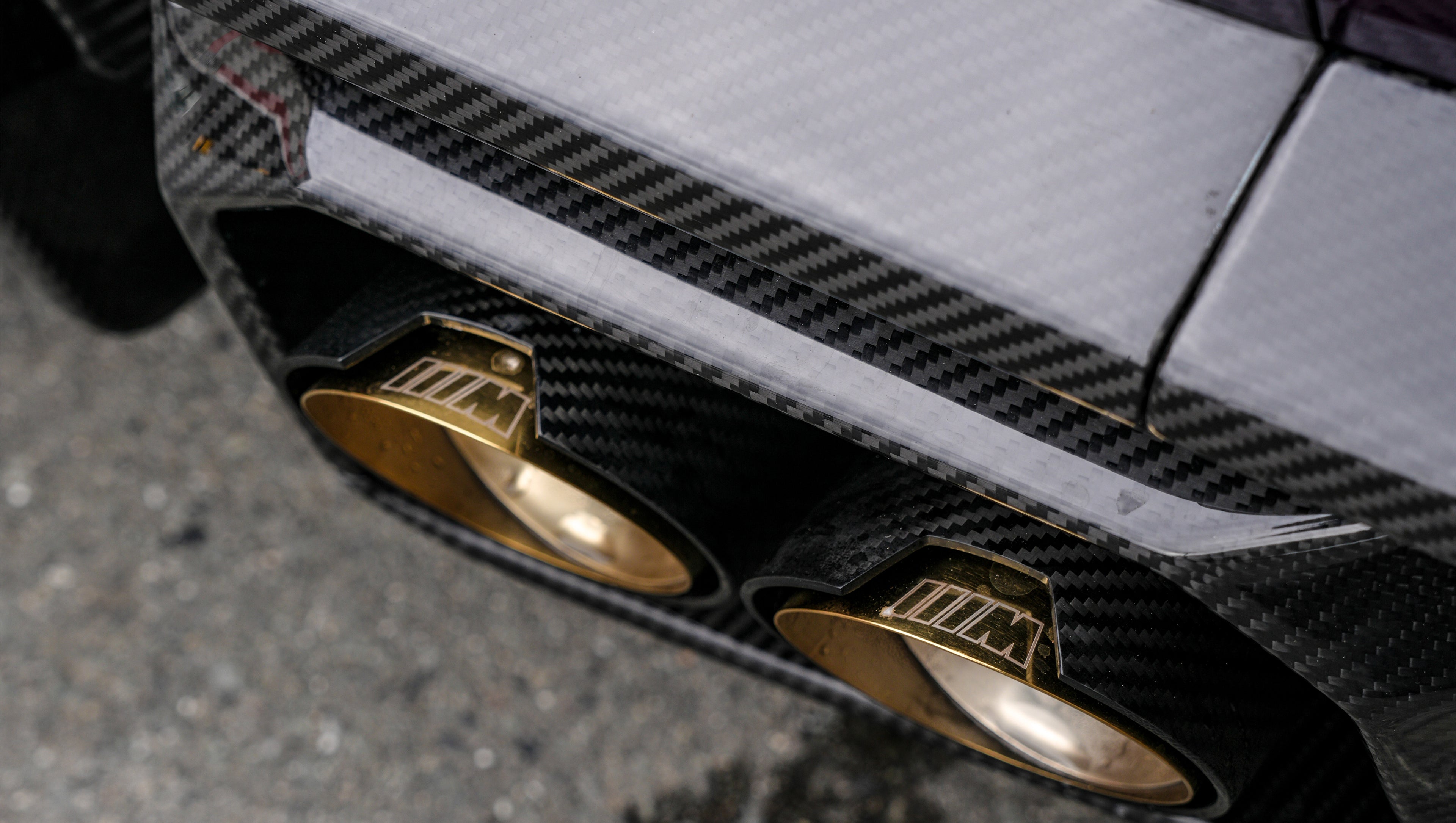 Close-up of REVOZPORT carbon fiber rear diffuser with dual gold exhaust tips, highlighting the glossy carbon weave and performance rear aero detail.