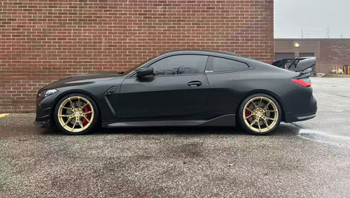 Side view of a matte black BMW G82 M4 featuring REVOZPORT carbon fiber side skirts, front lip, rear wing, and bronze forged wheels parked beside a brick wall.