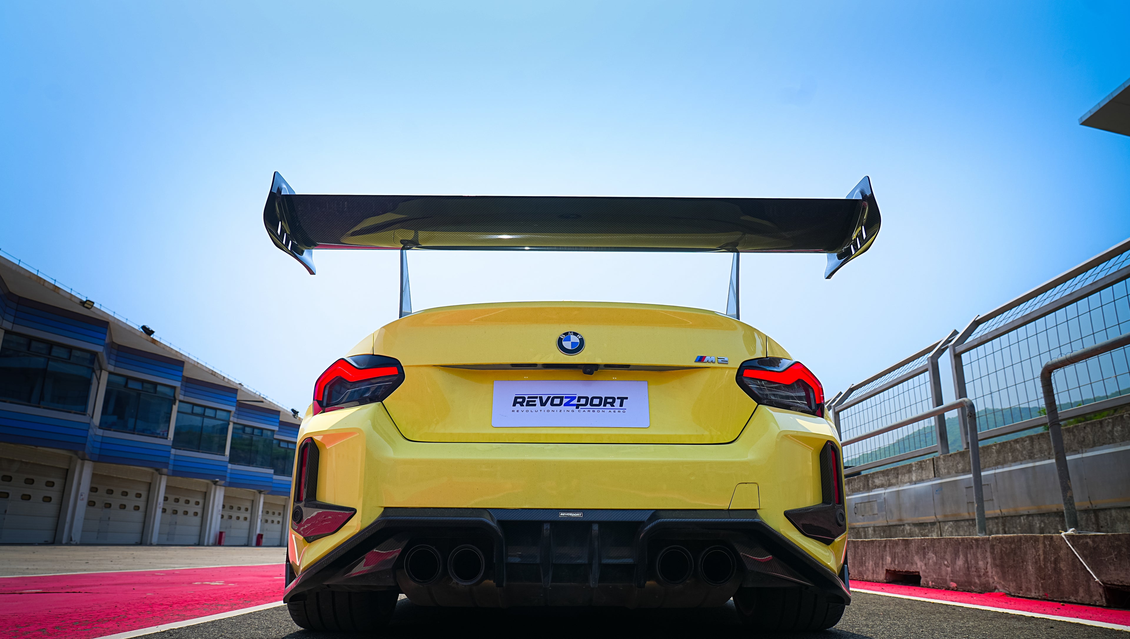 Yellow BMW G82 M4 with aggressive carbon fiber front splitter, vented hood, side canards, and bronze wheels parked on a race track pit lane.