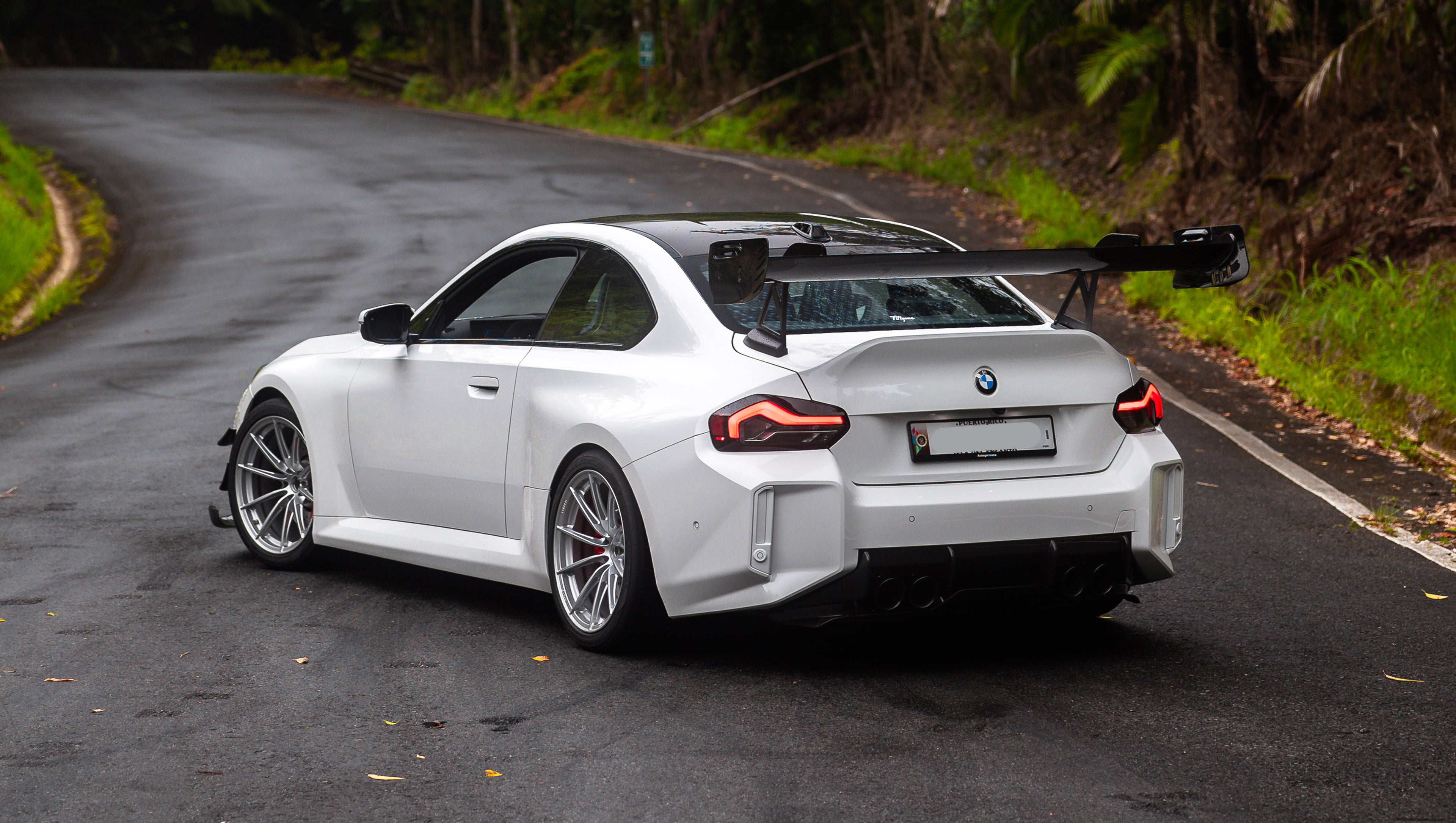 White widebody BMW G82 M4 with aggressive front bumper, canards, deep front splitter, large rear wing, and yellow LED headlights parked on a wet road surrounded by dense tropical forest.