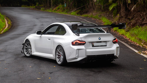 White widebody BMW G82 M4 with aggressive front bumper, canards, deep front splitter, large rear wing, and yellow LED headlights parked on a wet road surrounded by dense tropical forest.