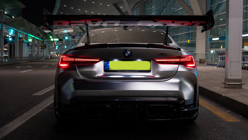 Matte gray BMW G82 M4 featuring an aggressive REVOZPORT carbon fiber aero kit with vented hood, front canards, wide fender vents, and illuminated LED headlights in a nighttime parking garage.