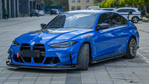 Blue BMW G80 M3 with REVOZPORT carbon fiber front splitter, side skirts, vented fenders, and large rear wing parked in a landscaped outdoor plaza.