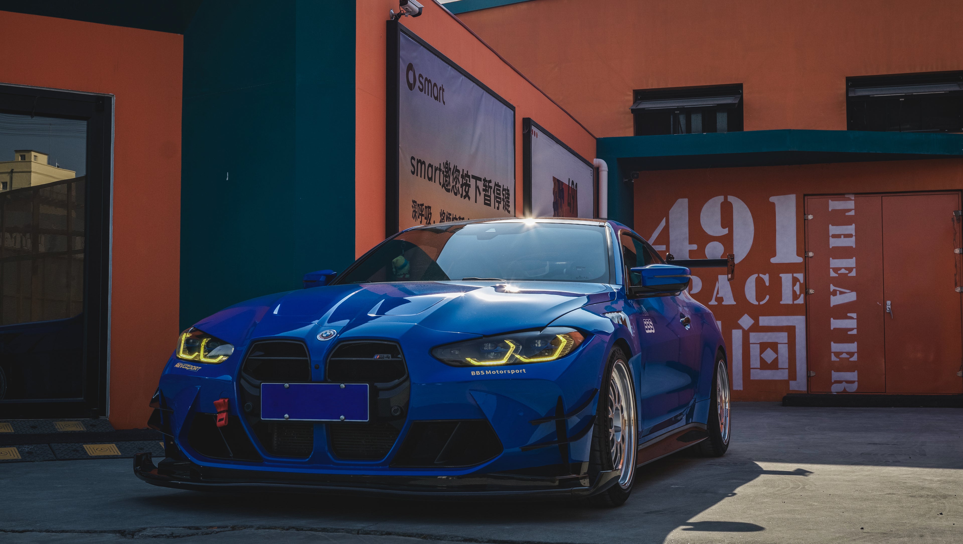 Blue BMW G82 M4 with carbon fiber front splitter, side skirts, rear wing, and aftermarket wheels parked in a modern industrial courtyard with colorful architectural walls.
