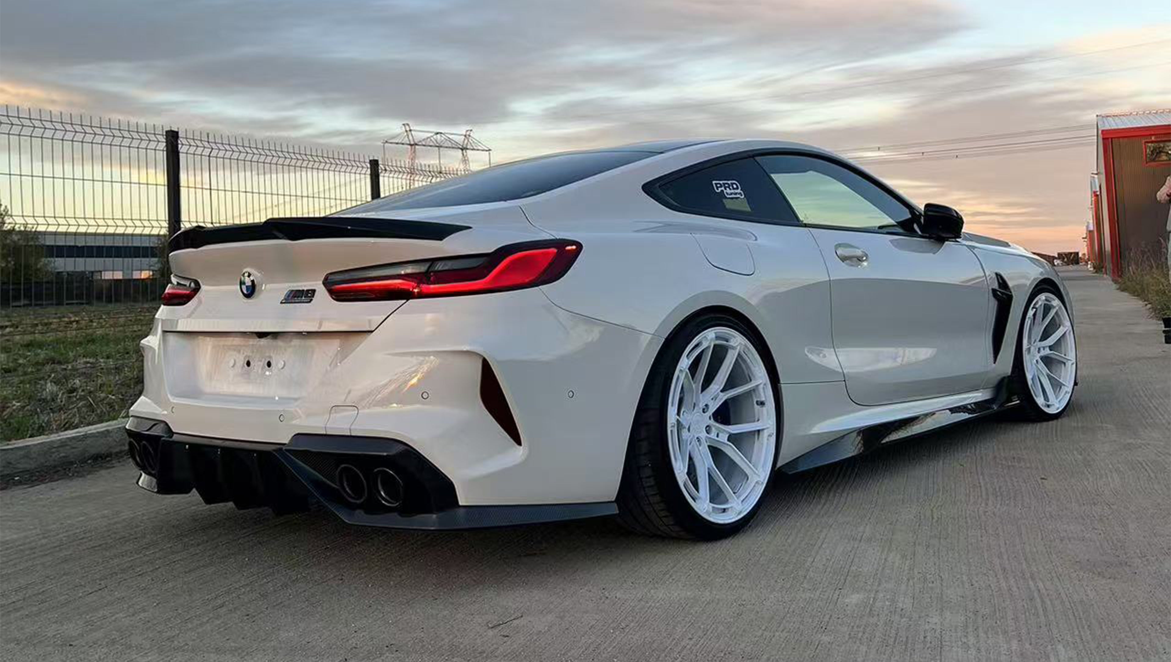 White BMW F92 M8 featuring a REVOZPORT carbon fiber front lip, side skirts, black front grille, and white multi-spoke forged wheels parked at sunset on an open road.