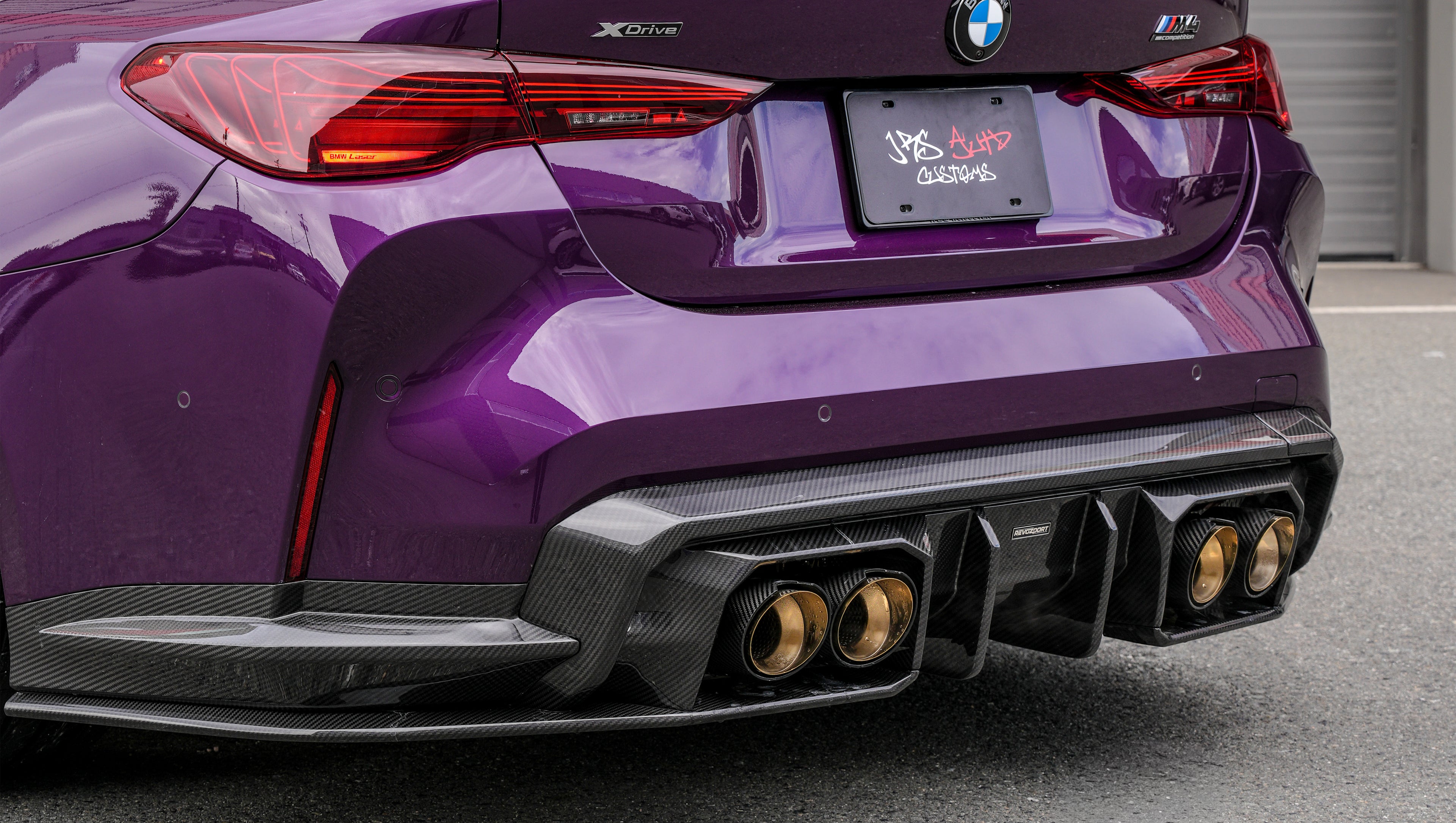 Purple BMW G82 M4 featuring a REVOZPORT carbon fiber body kit with front lip, side skirts, vented fenders, and gold wheels parked outside a modern industrial building.