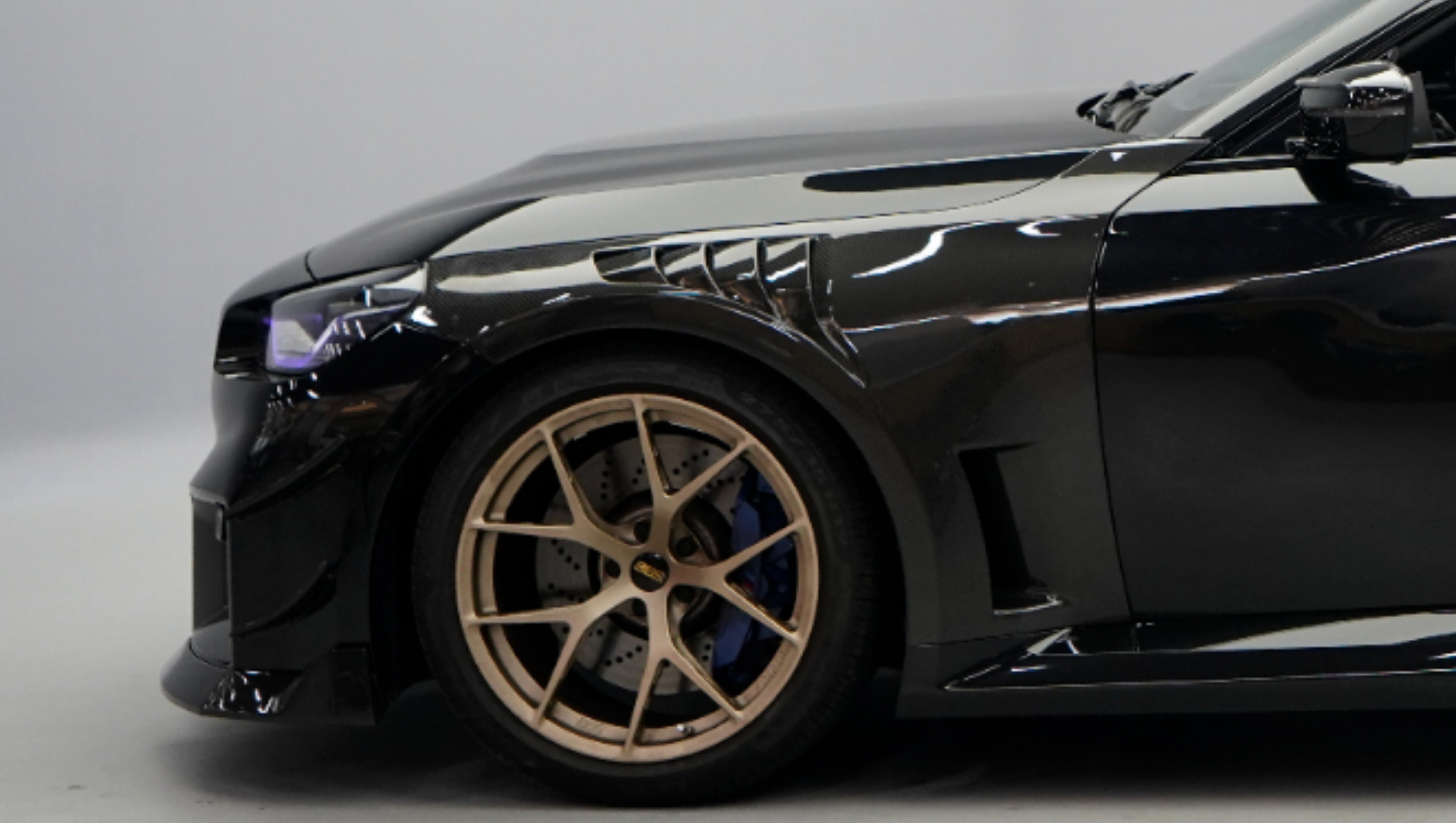 Front close-up of a black BMW G87 M2 featuring an aggressive carbon fiber front bumper, deep front splitter, large air intakes, and gloss carbon fiber aero details in a studio setting.
