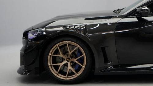 Front close-up of a black BMW G87 M2 featuring an aggressive carbon fiber front bumper, deep front splitter, large air intakes, and gloss carbon fiber aero details in a studio setting.