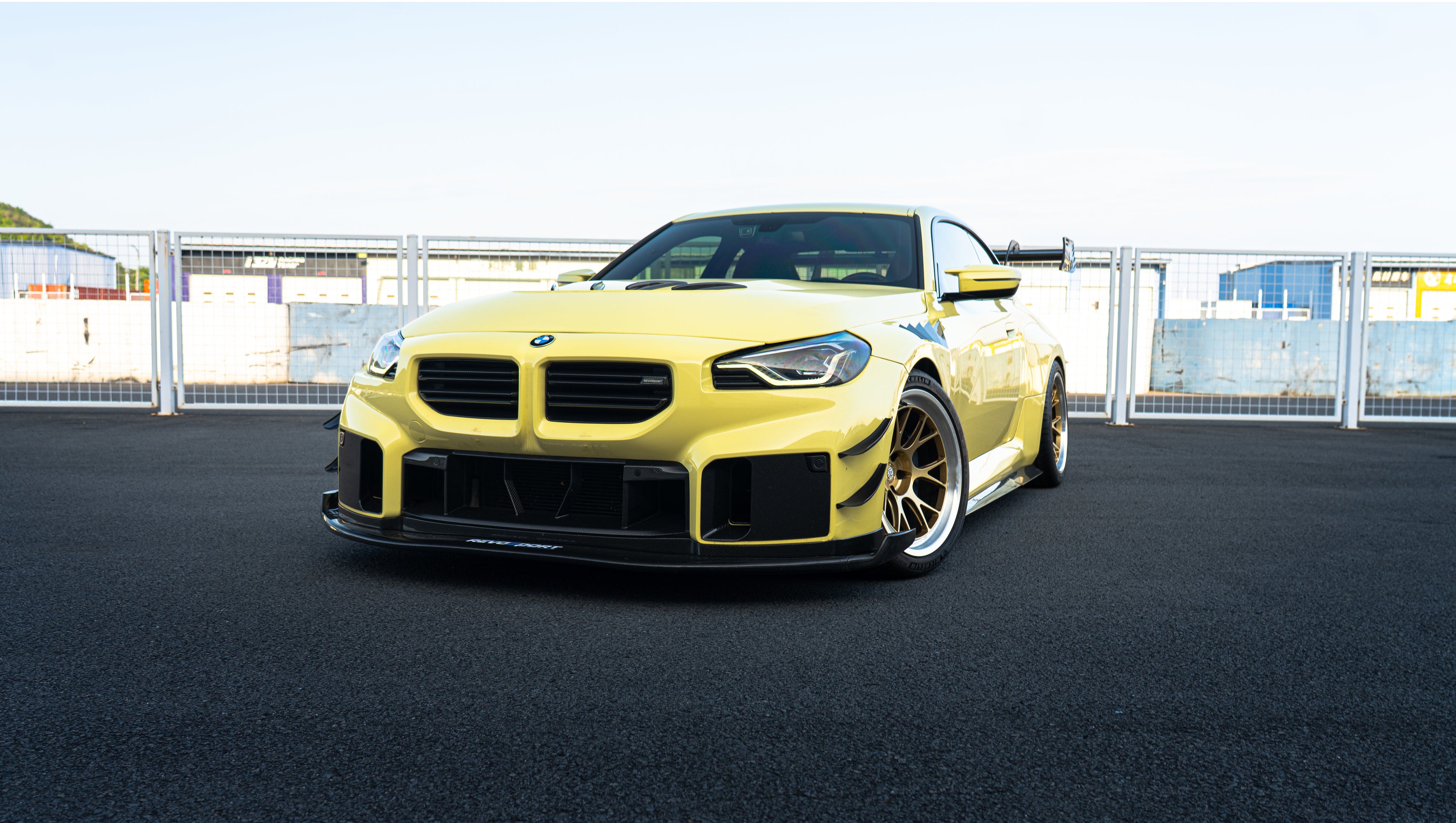 Pastel yellow BMW G82 M4 with carbon fiber front splitter, hood vents, side canards, bronze wheels, and large rear wing parked at a race track facility during golden hour.