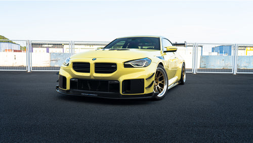 Pastel yellow BMW G82 M4 with carbon fiber front splitter, hood vents, side canards, bronze wheels, and large rear wing parked at a race track facility during golden hour.