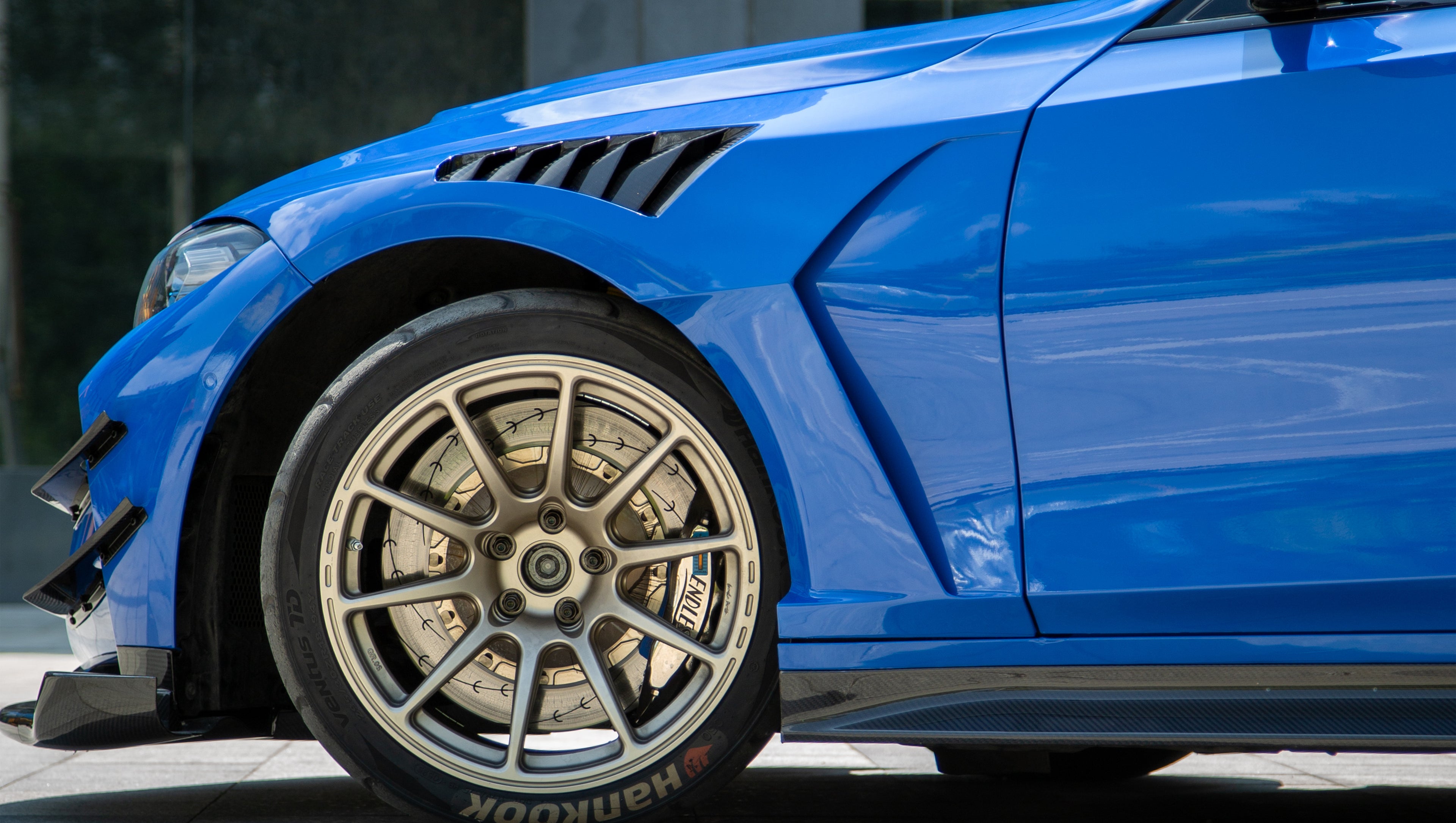 Blue BMW G80 M3 with REVOZPORT carbon fiber front splitter, side skirts, vented fenders, and large rear wing parked in a landscaped outdoor plaza.