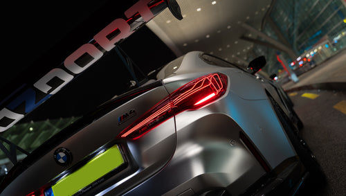 Matte gray BMW G82 M4 featuring an aggressive REVOZPORT carbon fiber aero kit with vented hood, front canards, wide fender vents, and illuminated LED headlights in a nighttime parking garage.