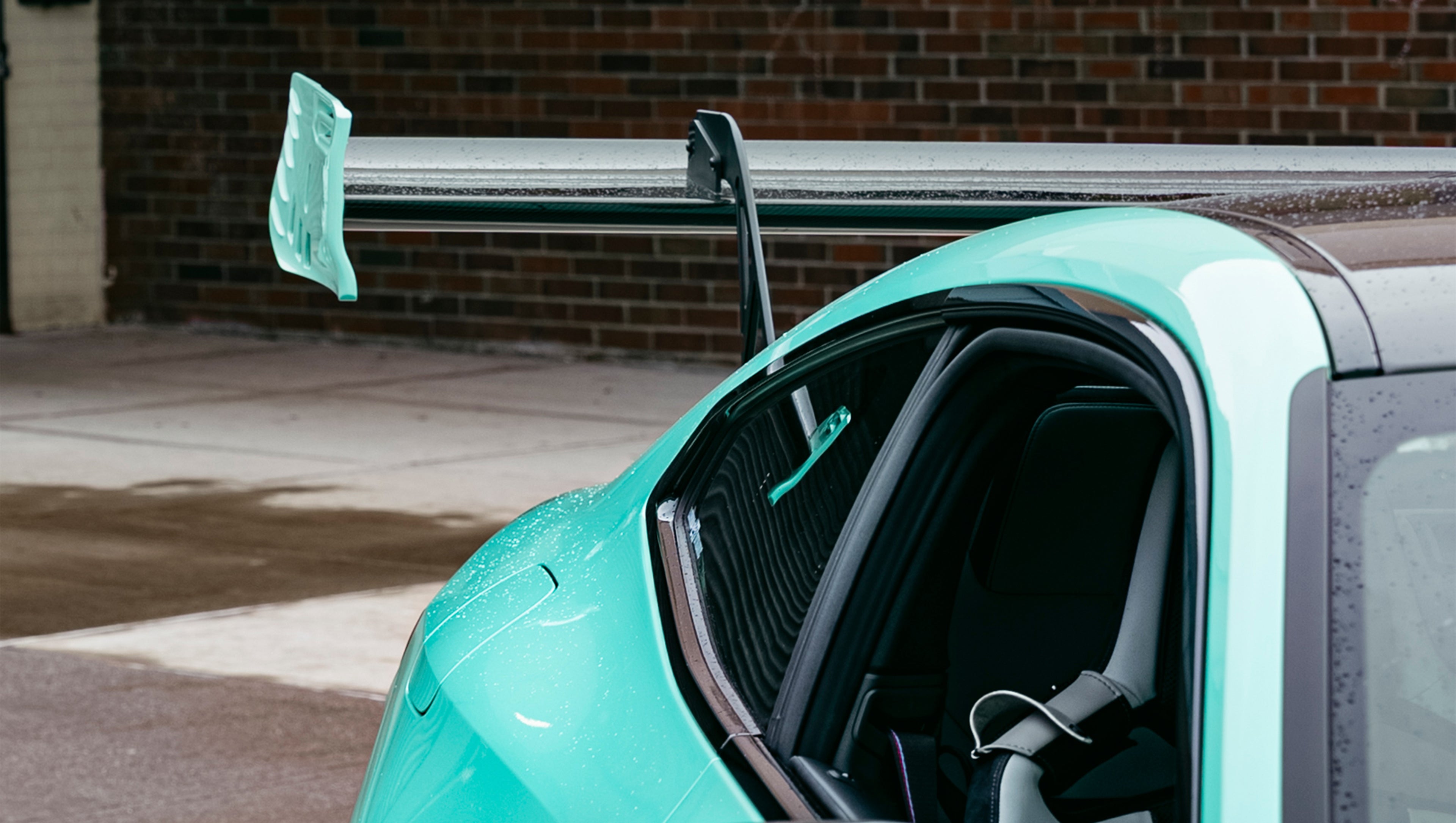 Mint green BMW G82 M4 with carbon fiber front splitter, side skirts, and large rear wing parked on a wet urban street outside a modern building.