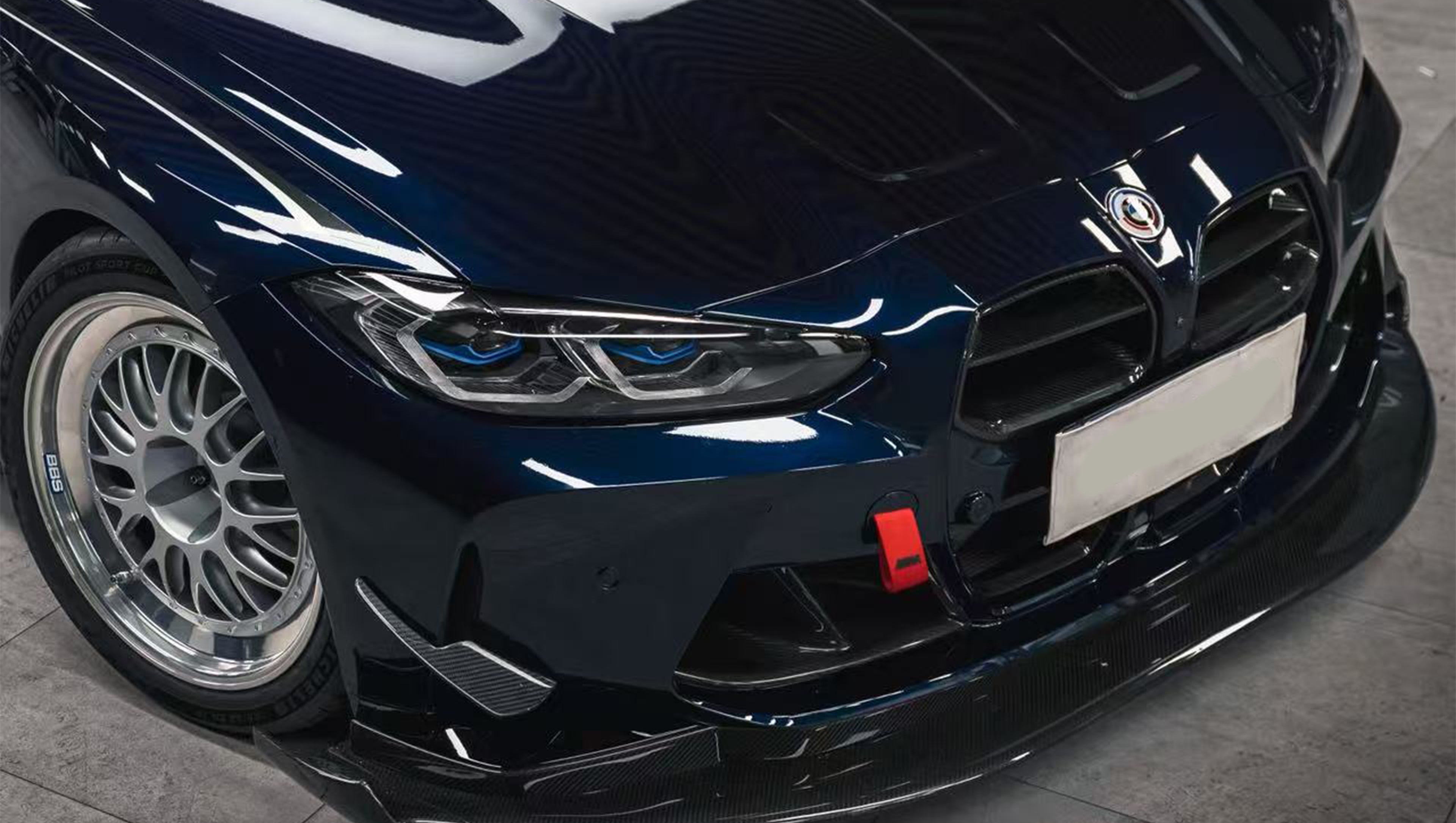 Dark blue BMW G82 M4 equipped with carbon fiber front splitter, side skirts, vented fenders, and a large rear wing, parked inside a modern luxury garage.