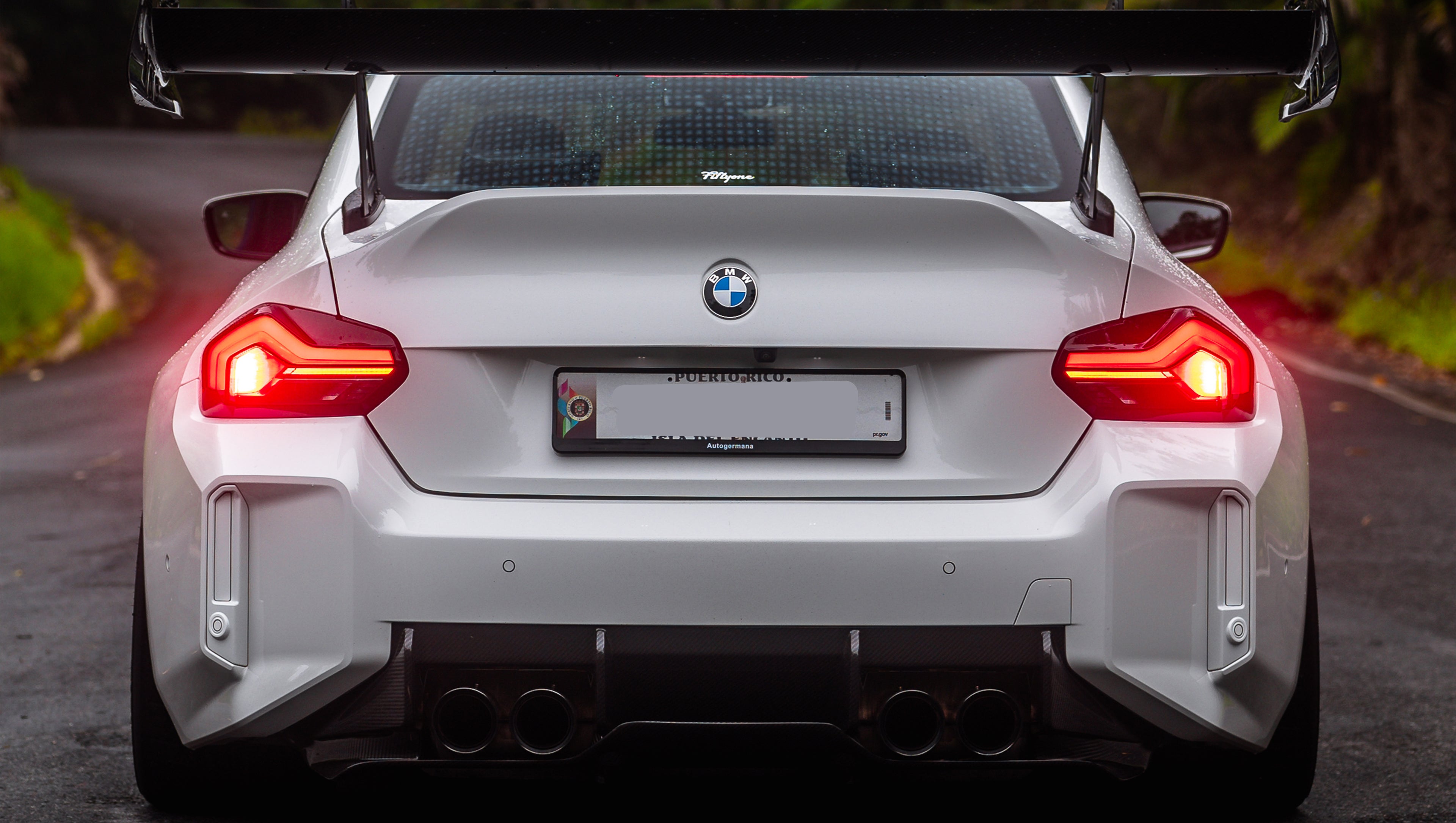 White widebody BMW G82 M4 with aggressive front bumper, canards, deep front splitter, large rear wing, and yellow LED headlights parked on a wet road surrounded by dense tropical forest.