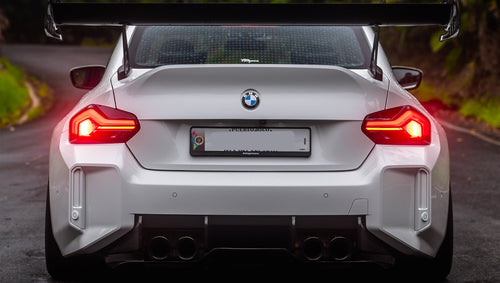 White widebody BMW G82 M4 with aggressive front bumper, canards, deep front splitter, large rear wing, and yellow LED headlights parked on a wet road surrounded by dense tropical forest.