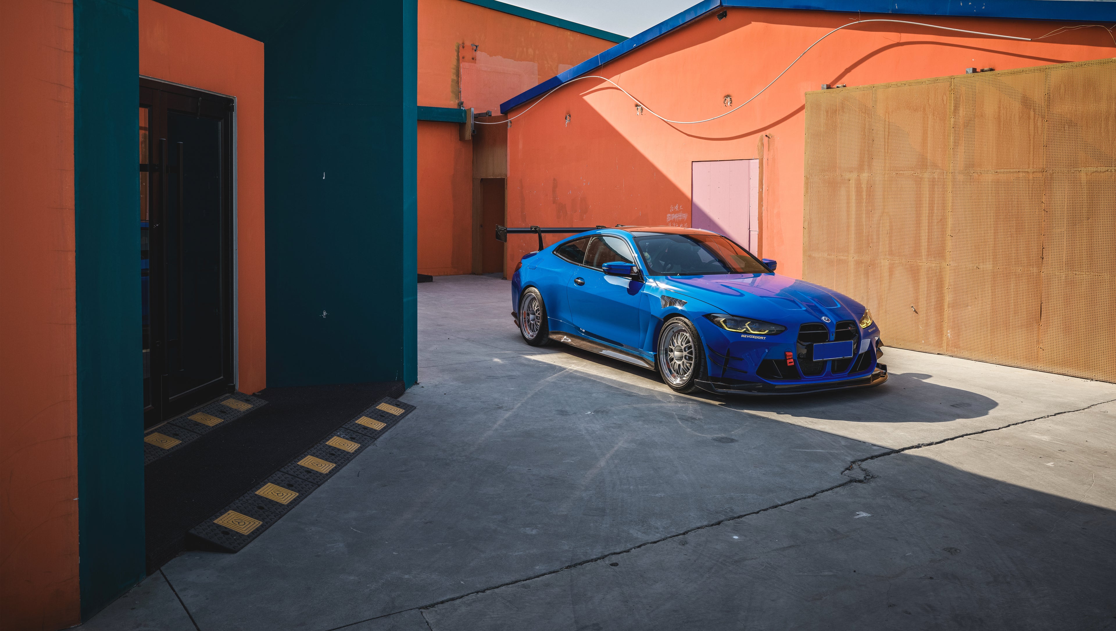 Blue BMW G82 M4 with carbon fiber front splitter, side skirts, rear wing, and aftermarket wheels parked in a modern industrial courtyard with colorful architectural walls.