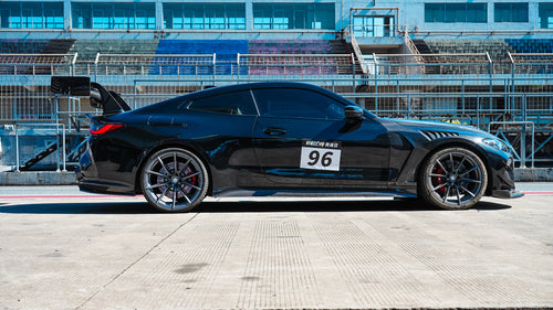 Black BMW G82 M4 track build with carbon fiber vented fenders, side skirts, rear diffuser, and oversized GT wing parked at a professional racing circuit pit lane.