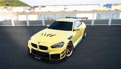 Pastel yellow BMW G82 M4 with carbon fiber front splitter, hood vents, side canards, bronze wheels, and large rear wing parked at a race track facility during golden hour.