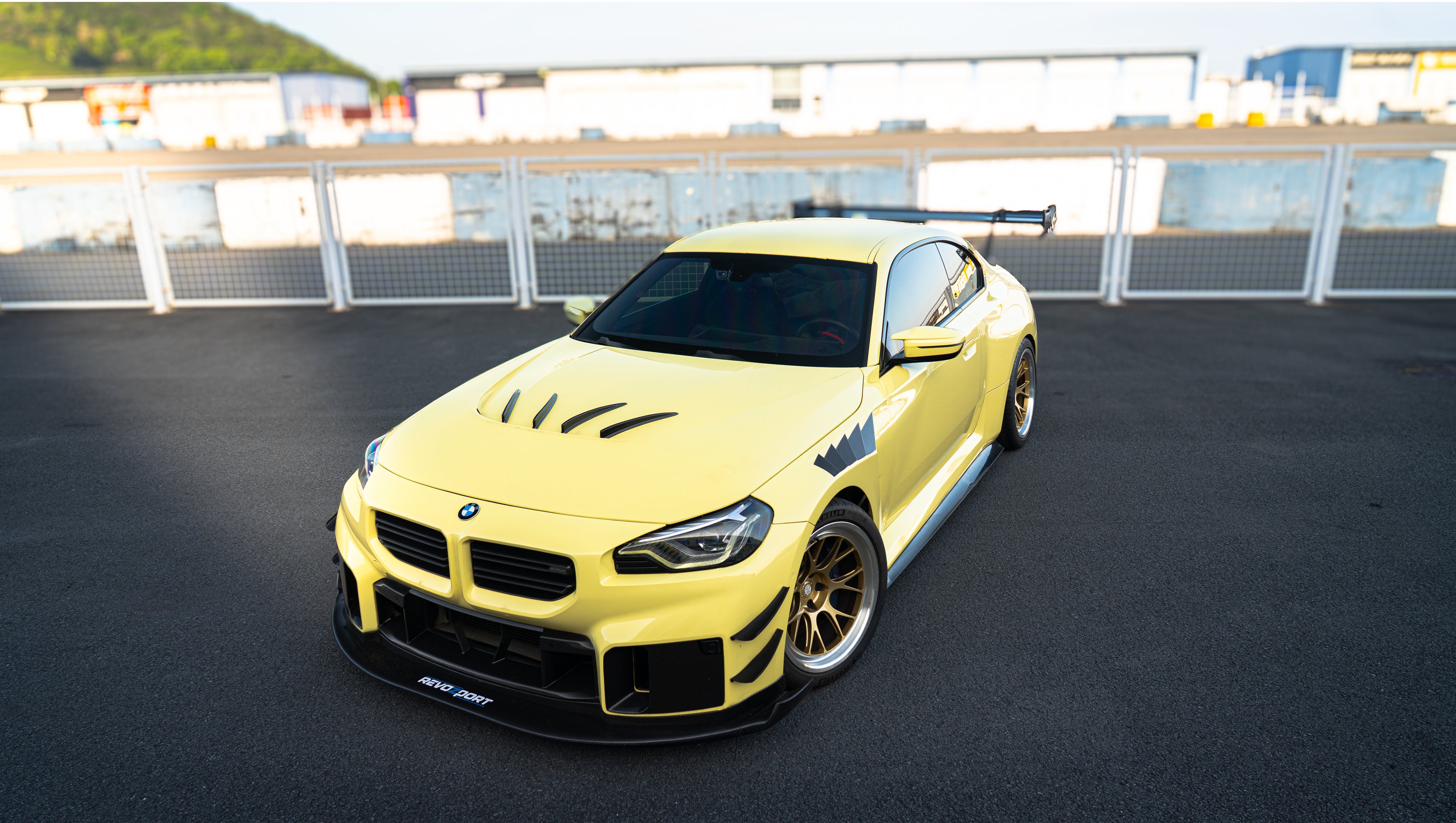 Pastel yellow BMW G82 M4 with carbon fiber front splitter, hood vents, side canards, bronze wheels, and large rear wing parked at a race track facility during golden hour.