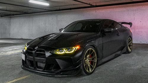 Side view of a matte black BMW G82 M4 featuring REVOZPORT carbon fiber side skirts, front lip, rear wing, and bronze forged wheels parked beside a brick wall.