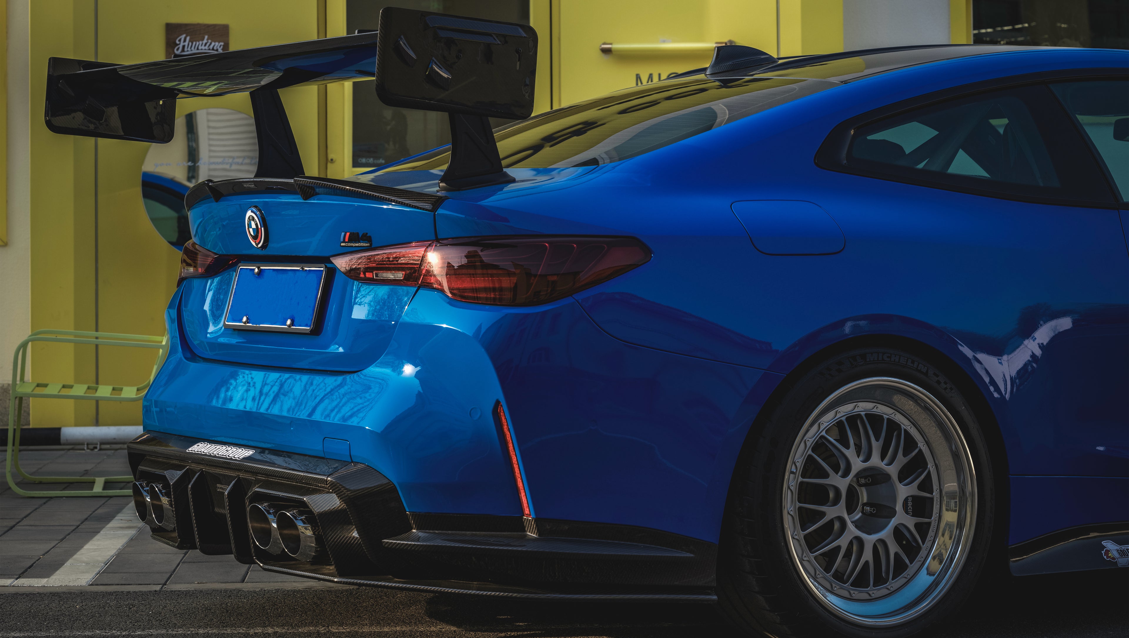 Blue BMW G82 M4 with carbon fiber front splitter, side skirts, rear wing, and aftermarket wheels parked in a modern industrial courtyard with colorful architectural walls.