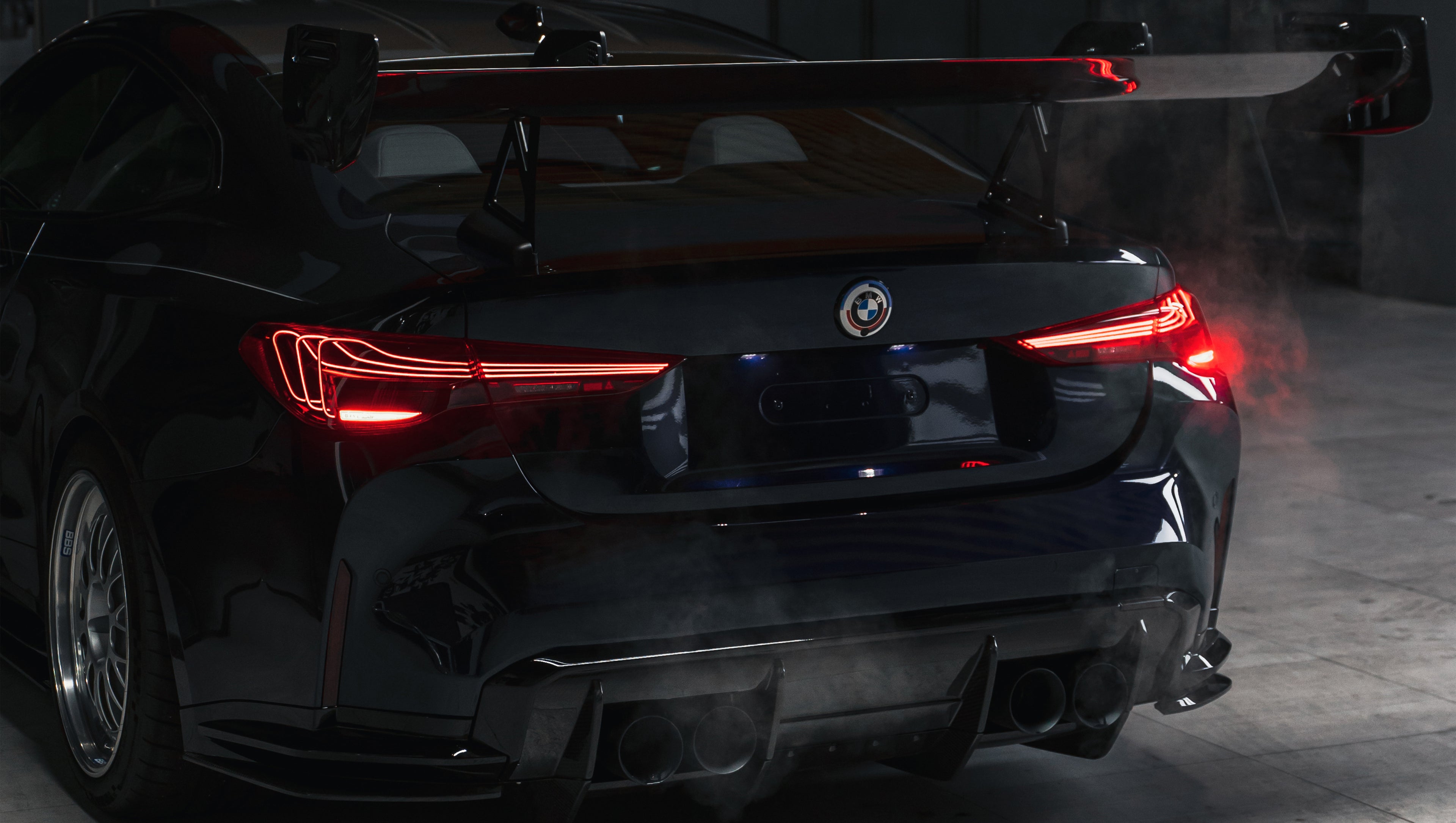 Dark blue BMW G82 M4 equipped with carbon fiber front splitter, side skirts, vented fenders, and a large rear wing, parked inside a modern luxury garage.