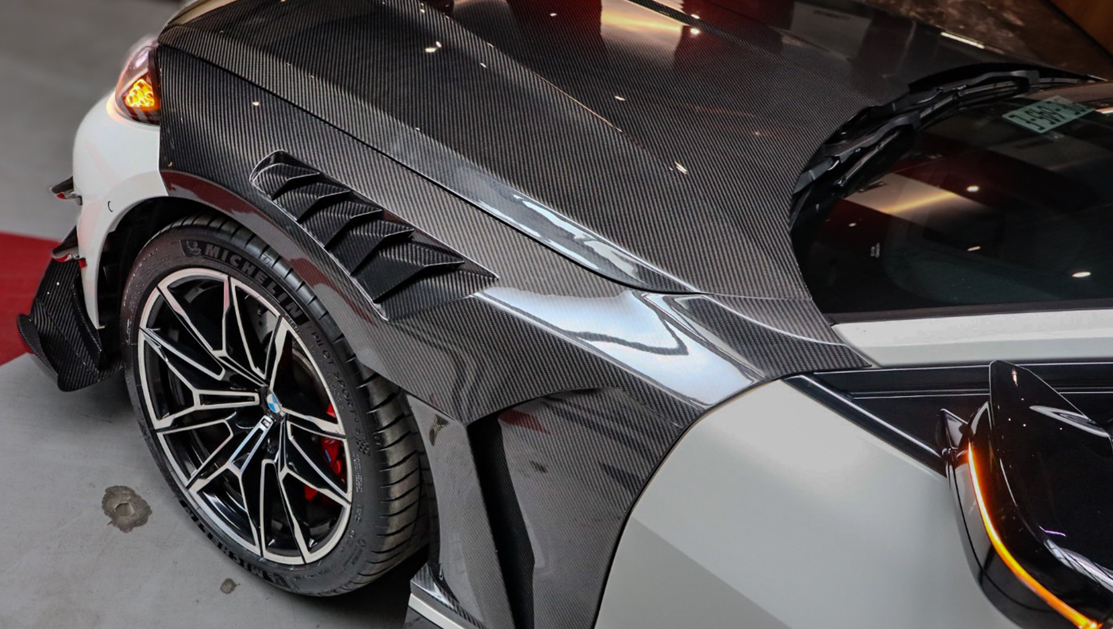White BMW G82 M4 featuring a forged carbon fiber hood, front splitter, side canards, and aggressive aero styling parked inside a modern underground garage.