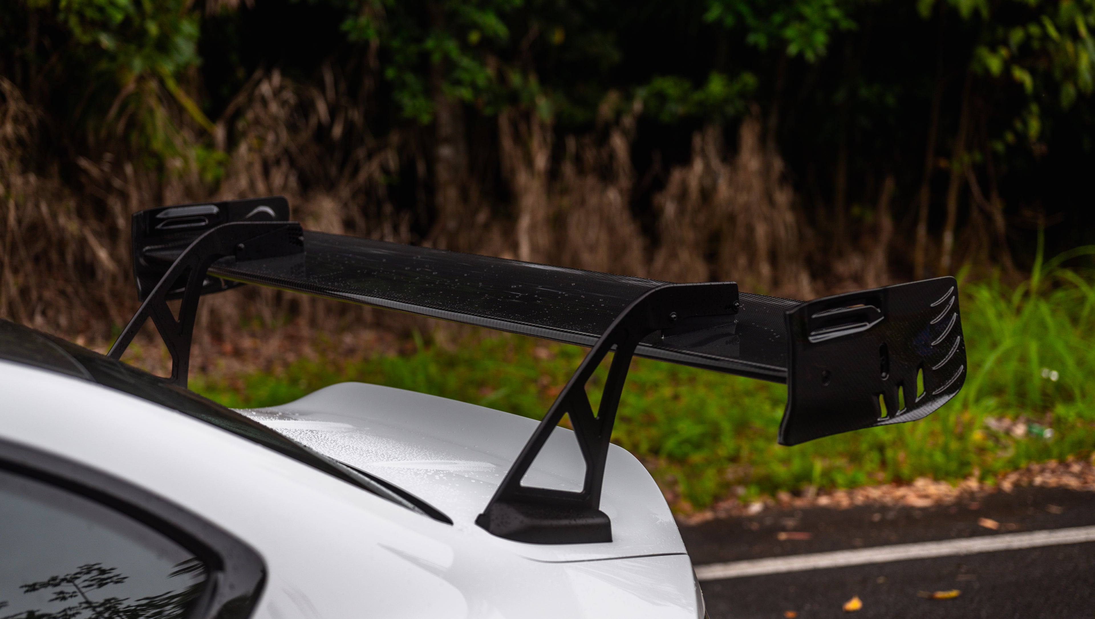 White widebody BMW G82 M4 with aggressive front bumper, canards, deep front splitter, large rear wing, and yellow LED headlights parked on a wet road surrounded by dense tropical forest.
