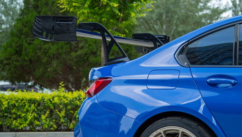 Blue BMW G80 M3 with REVOZPORT carbon fiber front splitter, side skirts, vented fenders, and large rear wing parked in a landscaped outdoor plaza.