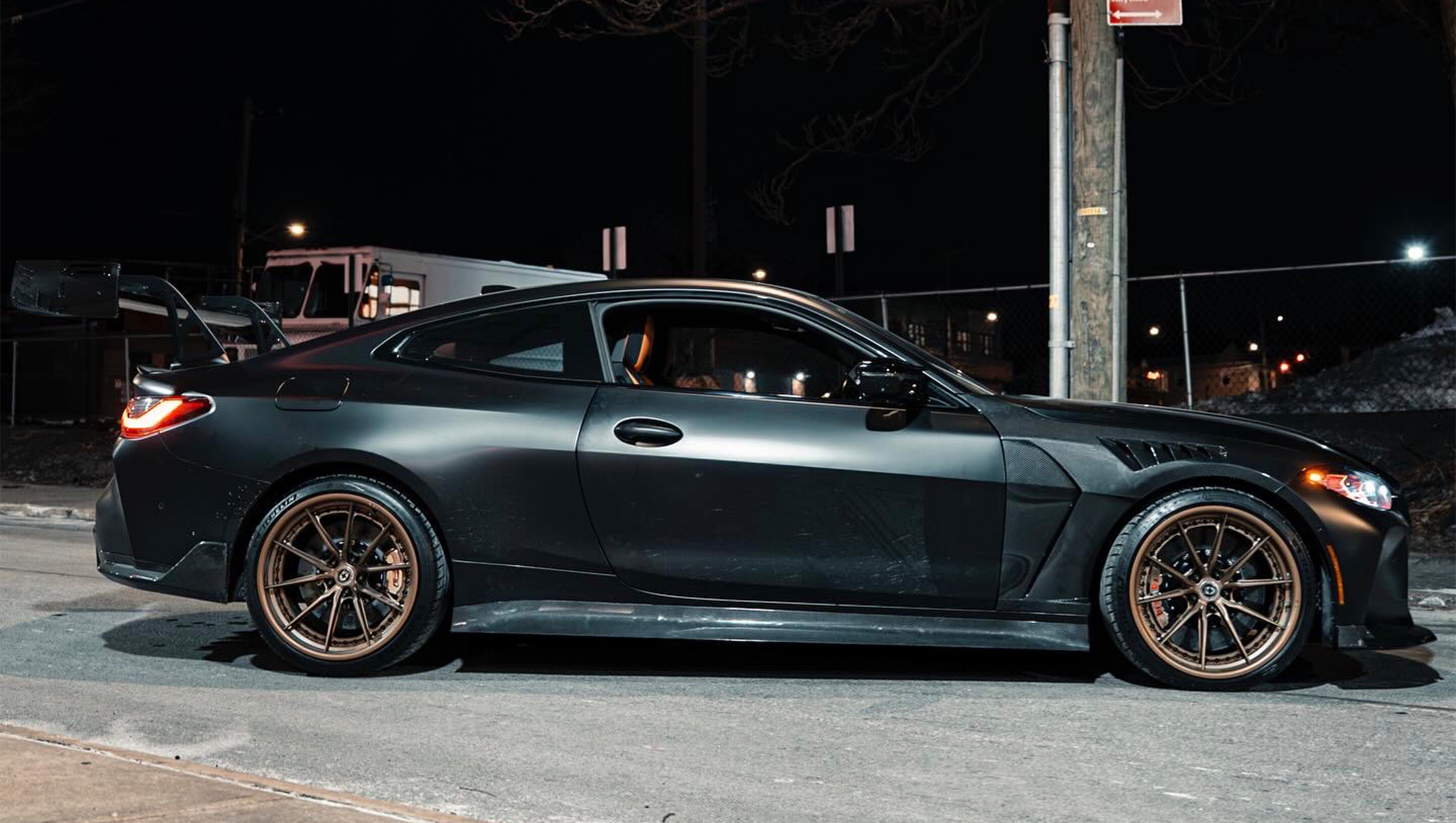 Matte black BMW G82 M4 with carbon fiber front splitter, side skirts, vented hood, bronze wheels, and oversized rear wing parked on a city street at night with headlights on.