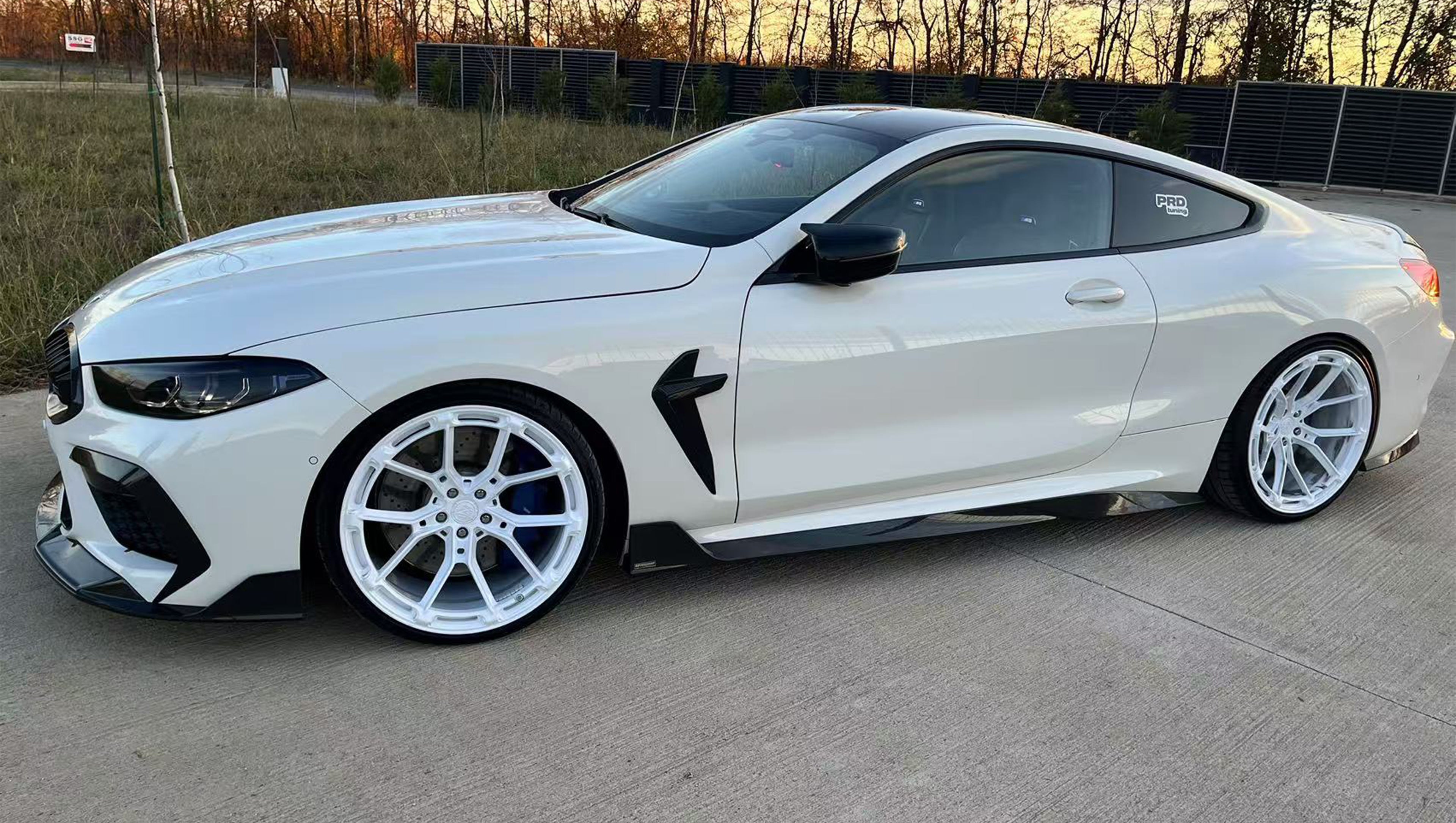 White BMW F92 M8 featuring a REVOZPORT carbon fiber front lip, side skirts, black front grille, and white multi-spoke forged wheels parked at sunset on an open road.