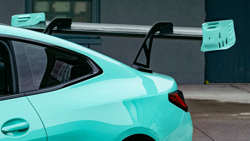 Mint green BMW G82 M4 with carbon fiber front splitter, side skirts, and large rear wing parked on a wet urban street outside a modern building.