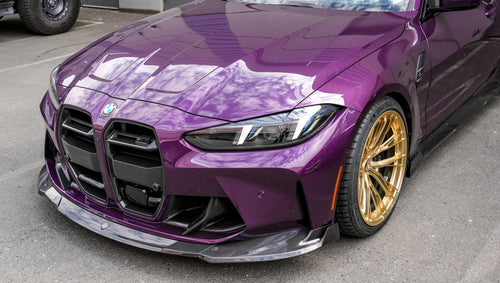 Purple BMW G82 M4 featuring a REVOZPORT carbon fiber body kit with front lip, side skirts, vented fenders, and gold wheels parked outside a modern industrial building.