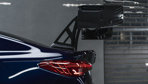 Dark blue BMW G82 M4 equipped with carbon fiber front splitter, side skirts, vented fenders, and a large rear wing, parked inside a modern luxury garage.
