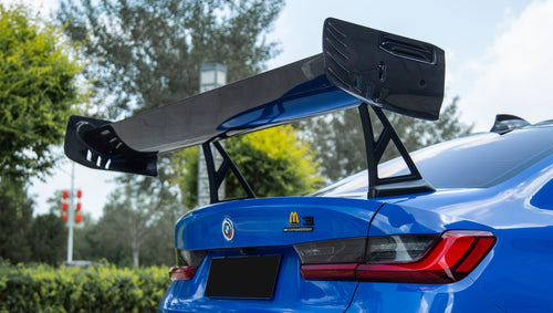 Blue BMW G80 M3 with REVOZPORT carbon fiber front splitter, side skirts, vented fenders, and large rear wing parked in a landscaped outdoor plaza.