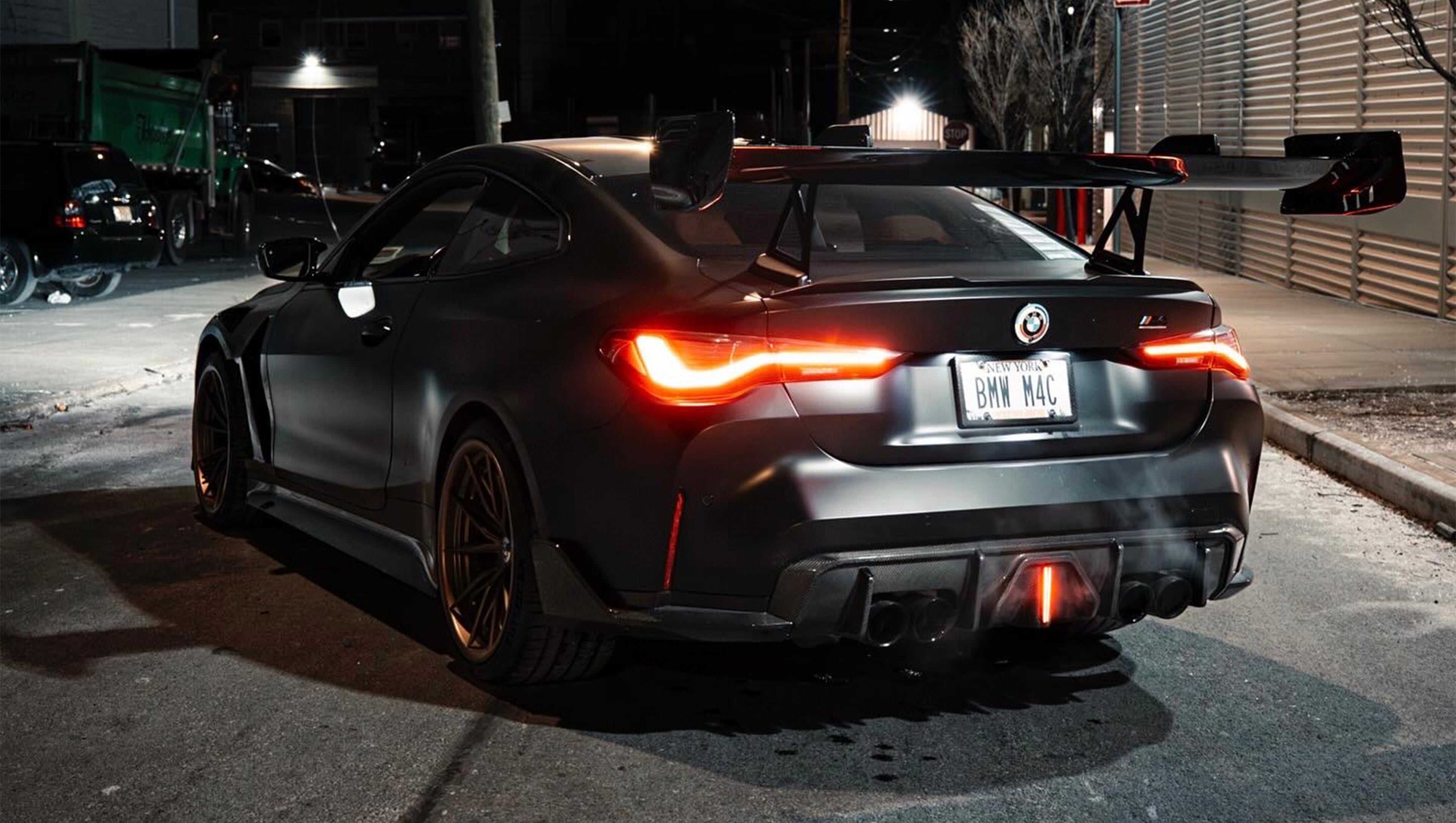 Matte black BMW G82 M4 with carbon fiber front splitter, side skirts, vented hood, bronze wheels, and oversized rear wing parked on a city street at night with headlights on.